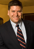 Headshot of a smiling male with dark hair, wearing a black blazer, white button down shirt, and red and black striped tie. He's pictured indoors.