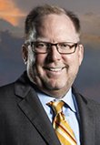 John Stewart. Caucasian male with light brown hair, wearing glasses and a suit coat with a light dress shirt and a yellow tie