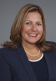 Paige Greenlee. Caucasian female with light brown shoulder-length hair wearing a navy suit jacket and white pearls.