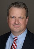 RJ Haughey. Caucasian male with light gray hair, wearing dark suit jacket, light blue dress shirt and red/white striped tie
