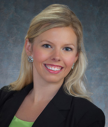 Melody Manning headshot, business woman with blonde hair is wearing a black suit and is pictured in front of a blue gray backdrop