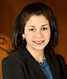Michelle Bedoya Barnett headshot, business woman with brown hair wearing a black suit with a blue shirt and necklace, pictured in front of a brown backdrop