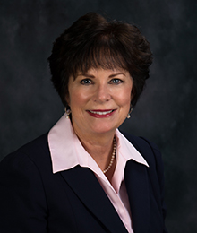 Michelle Jernigan headshot, Caucasian woman with short brown hair, wearing a black suit and pink button down shirt, pictured in front of a dark gray backdrop