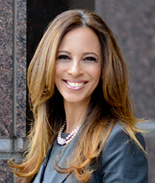 Michelle Suskauer headshot, young businesswoman smiling, she is photographed outdoors in front of a building, wearing a professional gray outfit and pearls