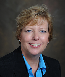 Patricia Crawels headshot, Caucasian business woman wearing a black suit with a light blue button down shirt pictured in front of a beige backdrop