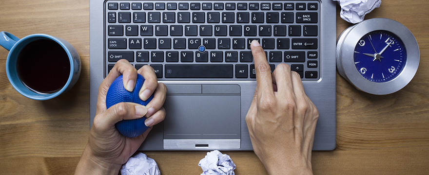 Close up of fingers typing on a laptop, one hand is holding a blue stress ball, a cup of coffee and crumpled papers surround the computer