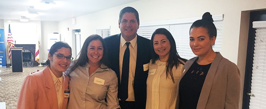 5 smiling business people wearing name tags, 4 women and 1 man, pose for a photo at a business meeting