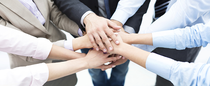 A group of people wearing business attire all holds their hands on top of one another to signify a unified team