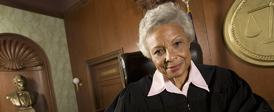 African American female judge with silver hair, smiling, wearing traditional judge clothing, seated on the bench in a courtroom setting