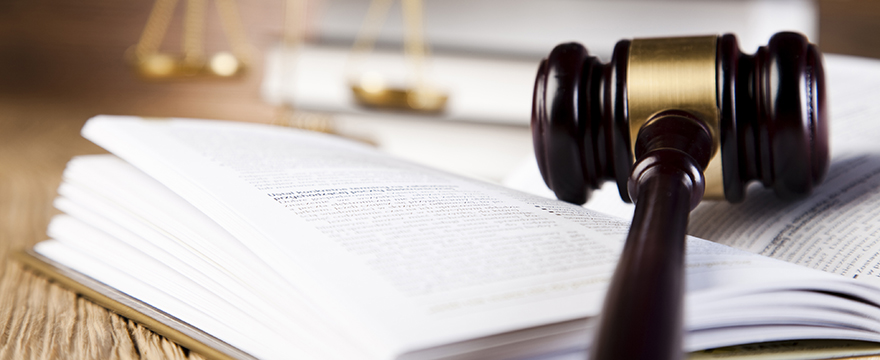Close up of a wood and gold gavel resting on an open page of a hardcover book, scales and another book are blurred in the background
