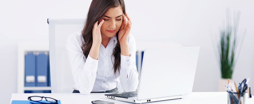 Young Caucasian female is rubbing her temples, looking down in a stressful stance. She's wearing a white button down shirt and sitting over a laptop at a desk