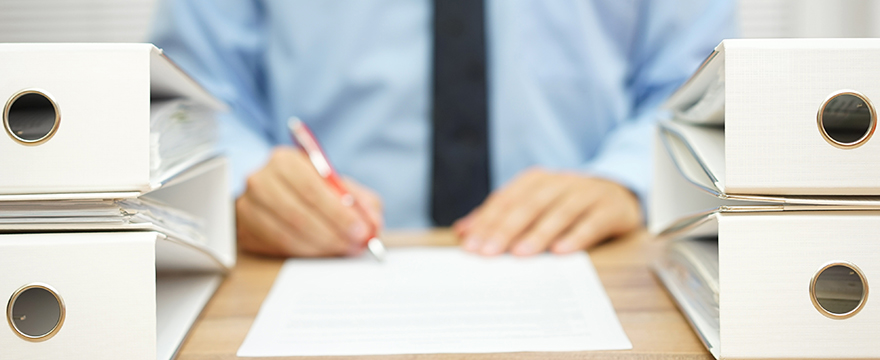 Straight on close up shot of a man in business attire, blue shirt and black tie. He's signing some paperwork with a red ink pen