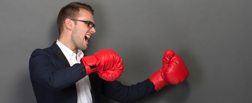 Young Caucasian male wearing a black suit is holding up his hands in a fighting stance, wearing red boxing gloves. His face is showing that he's yelling. Pictured in front of a gray backdrop.