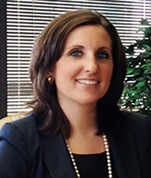Alexandria Hill, Caucasian female with brown hair, wearing a navy blazer and pearl necklace, pictured in an office setting in front of a window