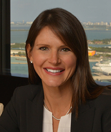 Deborah Baker Egozi headshot, Caucasian female with brown hair, wearing a white shirt under a black blazer, pictured in front of a window that overlooks a lake