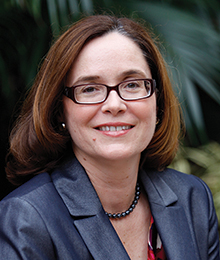 Debra Henley headshot, Caucasian woman photographed outdoors in front of a plant, she's wearing glasses and a navy suit