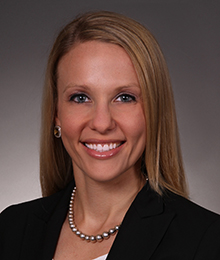 Elisa D'Amico headshot, Caucasian female with blonde hair wearing a black blazer and pearls, pictured in front of a beige backdrop