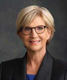 Elizabeth King headshot, Caucasian middle aged woman wearing glasses and a black suit, pictured in front of a solid gray background