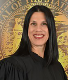 Honorable Laura Anne Stuzin, pictured in front of a large gold seal wearing a standard black academic judge gown