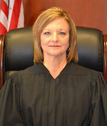 Honorable Margaret Hudson headshot, Caucasian woman with short light brown hair is pictured in a large leather chair with an American flag behind her