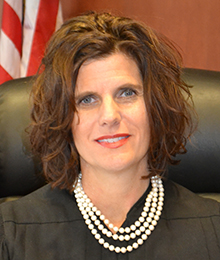 Honorable Sandra Upchurch headshot, Caucasian woman with short brown hair and a pearl necklace is pictured in a large leather chair with an American flag behind her