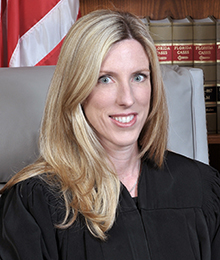 Honorable Stacy Ross headshot, middle aged Caucasian female with blonde hair is seated on a large office chair with law books and an American flag in the background