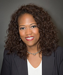 Kamilah Perry headshot, African American businesswoman with dark curly hair is wearing a black suit with a white shirt, pictured in front of a gray backdrop