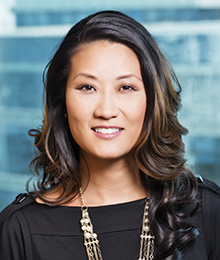 Katie Phang headshot, Asian businesswoman with long dark hair is wearing a black top with a statement necklace, pictured in front of a blue background