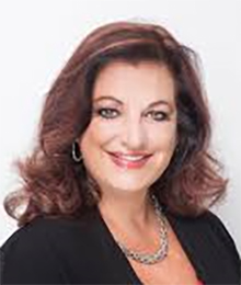 Mayanne Downs headshot, Caucasian woman dressed in a black business top, she has brown hair and is pictured in front of a white background
