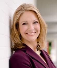 Melanie Griffin headshot, professional looking Caucasian woman leaning against a wall, smiling, she has blonde hair and is wearing a plum colored suit