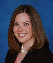 Miller, Caucasian female with brown hair wearing a black blazer and pearl necklace, pictured in front of a blue backdrop