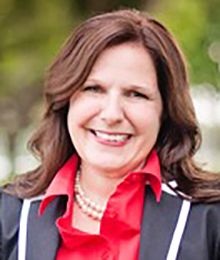 Paige Greenlee headshot, Caucasian female, middle aged with brown hair, wearing a black and white blazer with a red button down shirt and a pearl necklace