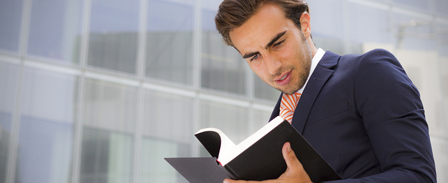 Young Caucasian male wearing a black suit is reading a black hard cover book in front of a gray concrete building