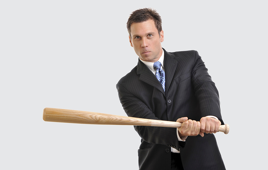 Businessman wearing a dark suit with a blue tie is holding a baseball bat in a ready to hit position, staged against a white background
