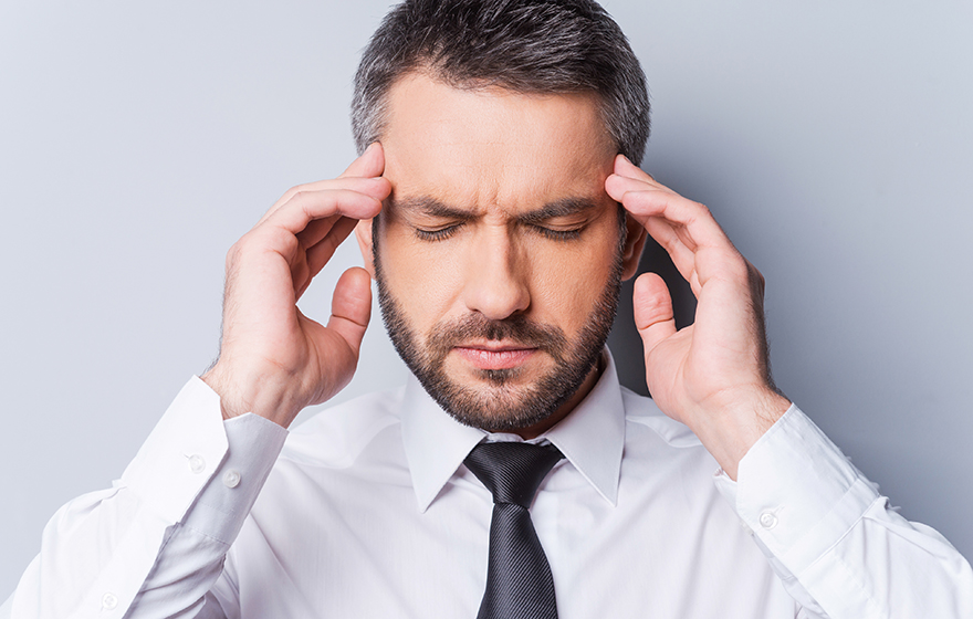 Stressed businessman has his hands on his temples, eyes closed, standing in front of a gray background