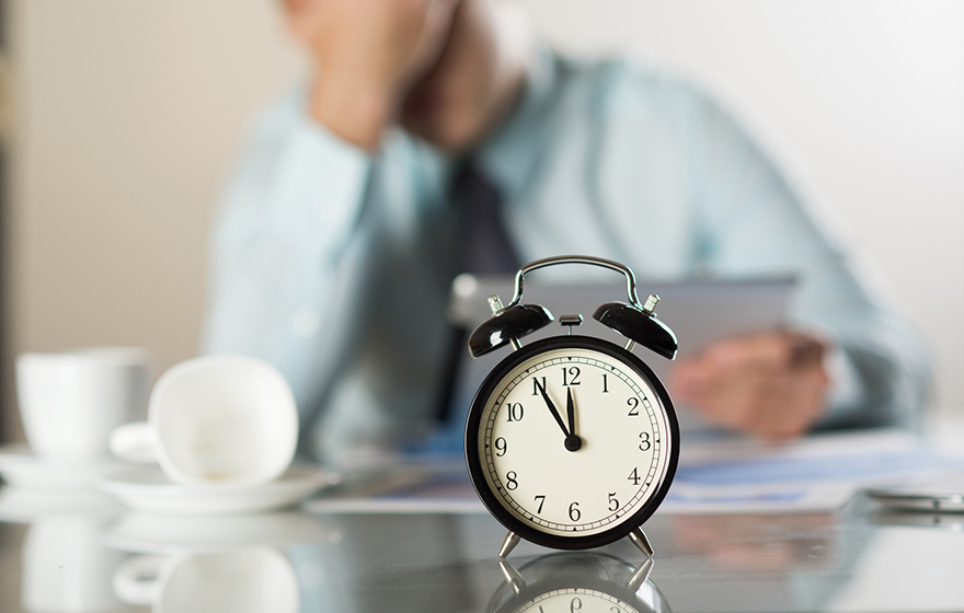 Small alarm clock in the foreground with a stressed businessman sitting at a desk in the background
