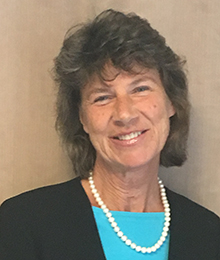 Carey Haughwout pictured in front of a brown background, wearing a black sweater, blue shirt and pearl necklace