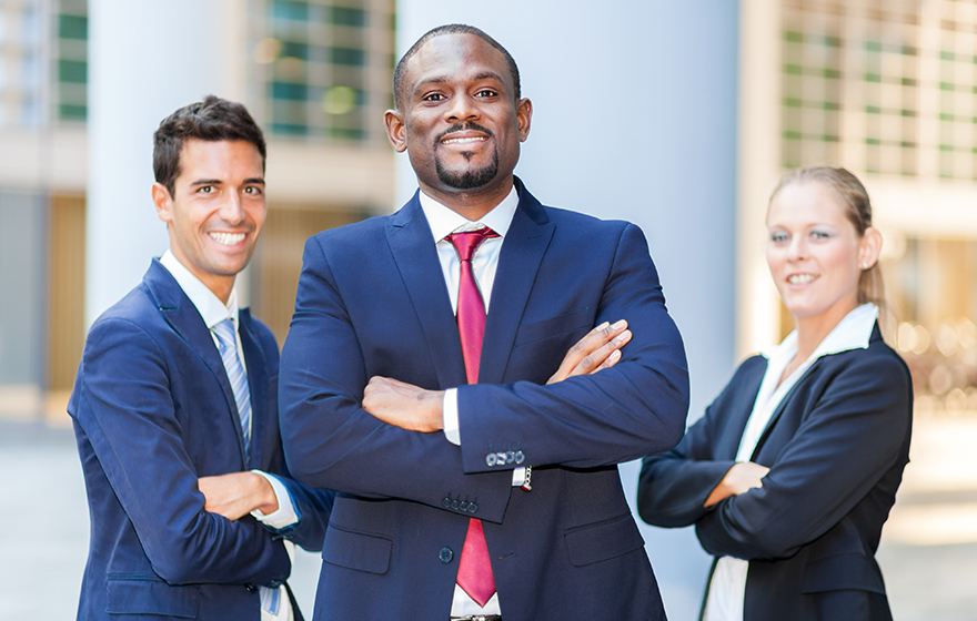 Group of business people, arms crossed, wearing navy suits, gentleman in front is African American with a red tie