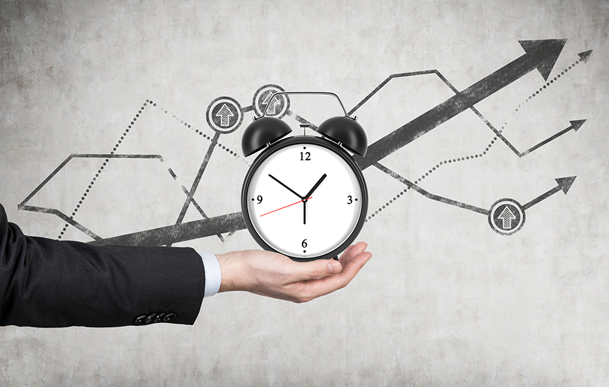 A businessman's hand holds an alarm clock. There is a growing line charts behind the alarm clock.