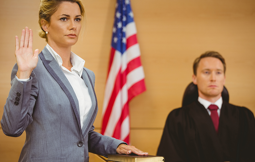 Female witness with hand on bible wearing gray suit with a white shirt, American flag and a judge pictured in the background