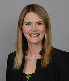 Caucasian young woman with light brown hair, silver necklace, black suit coat in front of a gray background