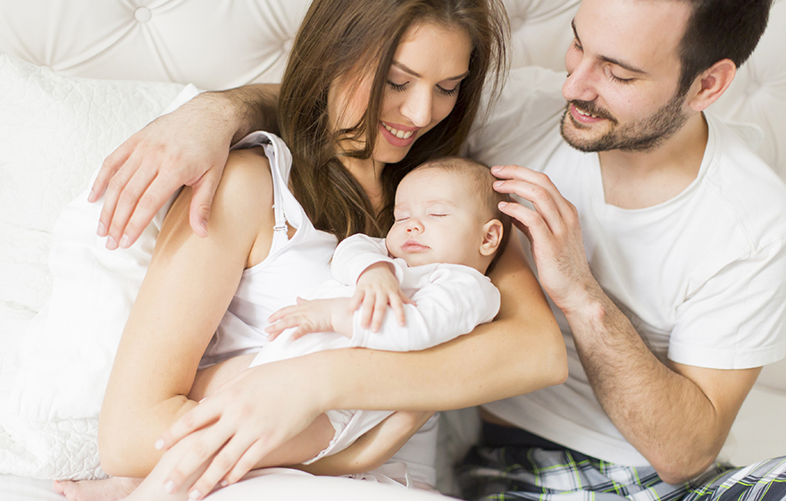 Proud mom and dad, with new baby snuggled together in bed. All wearing white pajamas in a white bed.