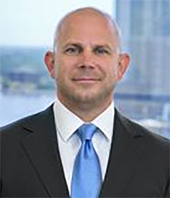Bill Adams headshot of a bald Caucasian man in a black suit, white shirt and bright blue tie. There is a building and water in the background