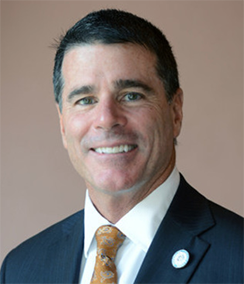 Bill Schifino headshot of a middle aged Caucasian man with dark brown hair wearing a navy blue suit, white shirt, and orange tie