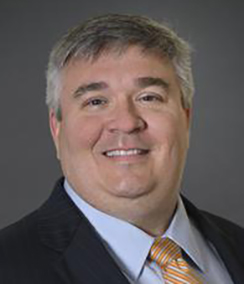 Brian Burgoon headshot of a middle aged smiling Caucasian man with grey hair, he is wearing a black suit with a light colored shirt and an orange striped tie