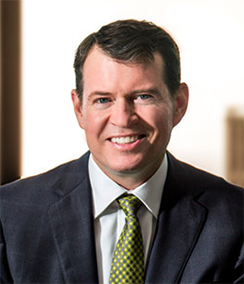 Bruce Denson headshot of a smiling middle aged Caucasian man with brown hair wearing a blue suit, white shirt and a green checkered tie