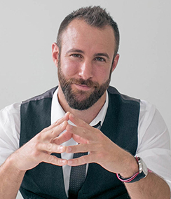 Ethan Wall headshot of a young Caucasian man with brown hair and a beard. He is smiling and has his hands up on a table.