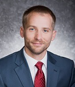 John Miller headshot of a young Caucasian man with light brown hair and beard in a navy blue suit, white shirt and red tie