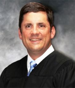 Judge F Rand Wallis headshot of a Caucasian man with brown hair and brown eyes in a black judges' robe with a white shirt and blue striped tie