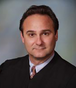 Judge Gary Flower headshot of a middle aged Caucasian man with brown hair and brown eyes in a black judges' robe. He also has a light colored shirt on a maroon tie.